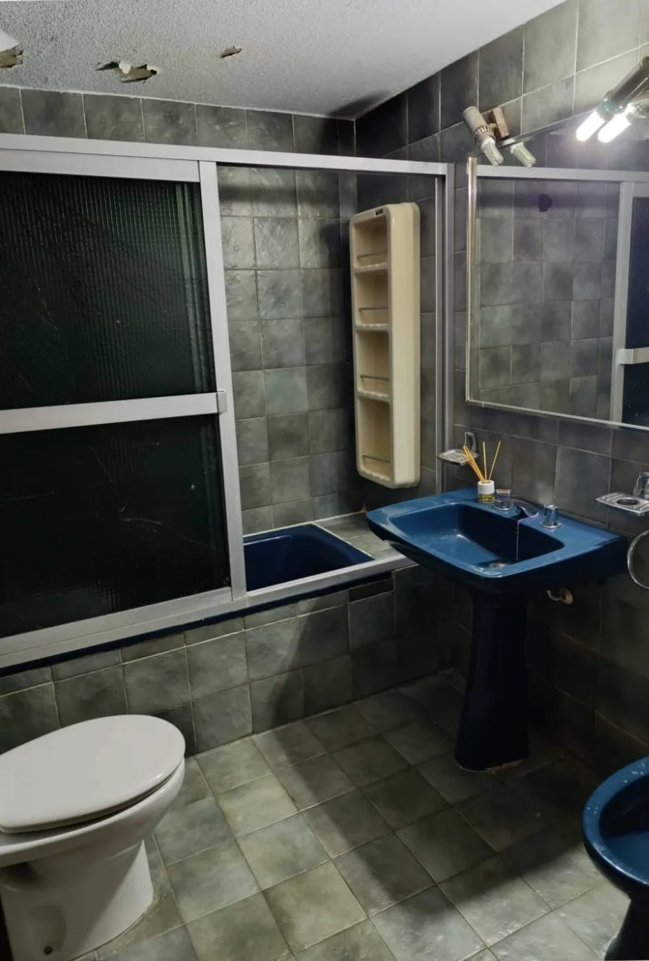 Baño antes de la reforma, con azulejo gris, bañera y lavabo de pie en azul y el techo desconchado