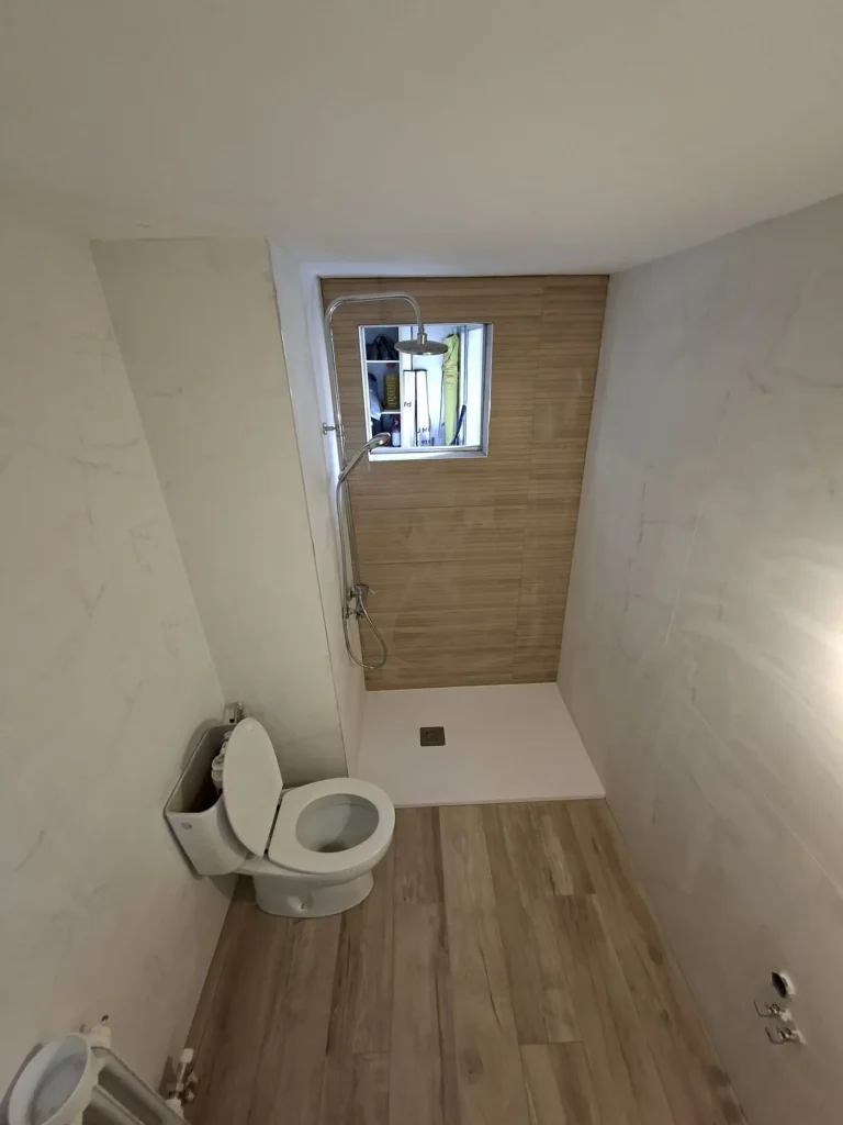 Baño reformado en la calle Joaquín Blumer (Móstoles), con ducha de plato blanco y pared efecto madera bajo la ventana