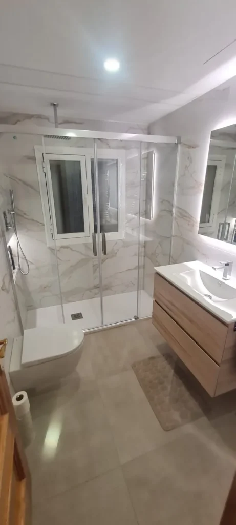 Baño reformado con plato de ducha enrasado, mampara corredera de cristal y paredes efecto mármol