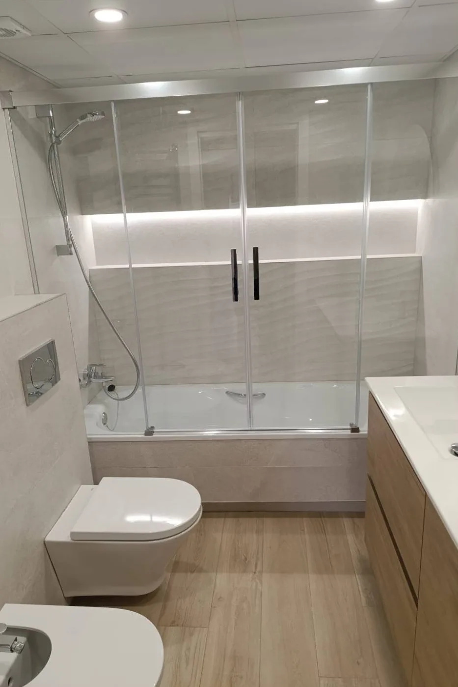 El mismo baño reformado, con bañera y mampara de dos hojas, hornacina con luz LED, inodoro suspendido y suelo efecto madera