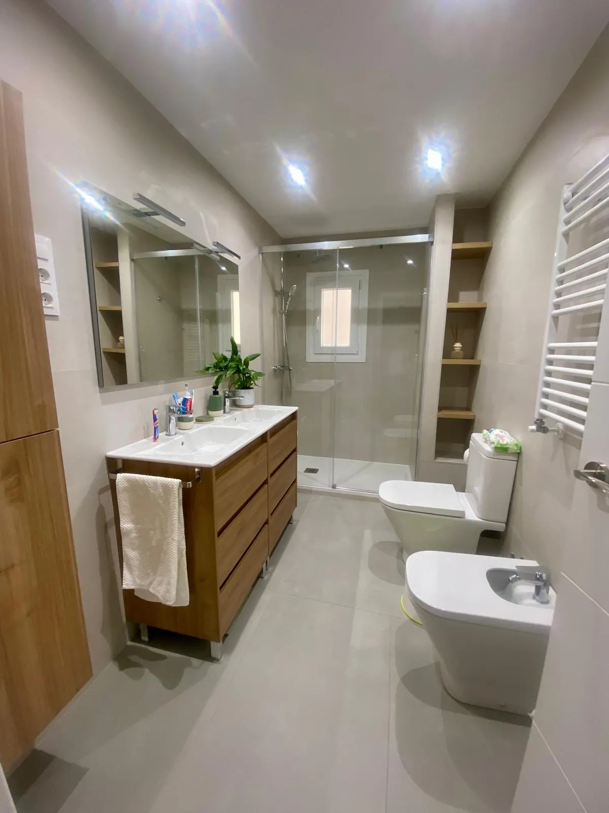 Baño reformado en Móstoles con ducha de cristal, estantes empotrados, mueble de lavabo de madera y radiador toallero