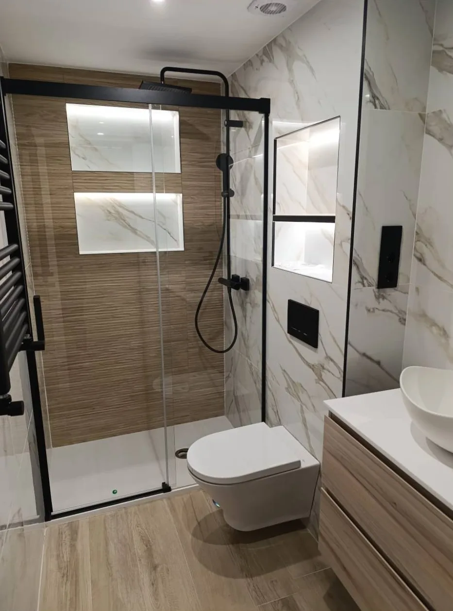 El mismo baño reformado, con ducha de mampara de perfil negro, pared efecto madera, hornacinas iluminadas y porcelánico efecto mármol