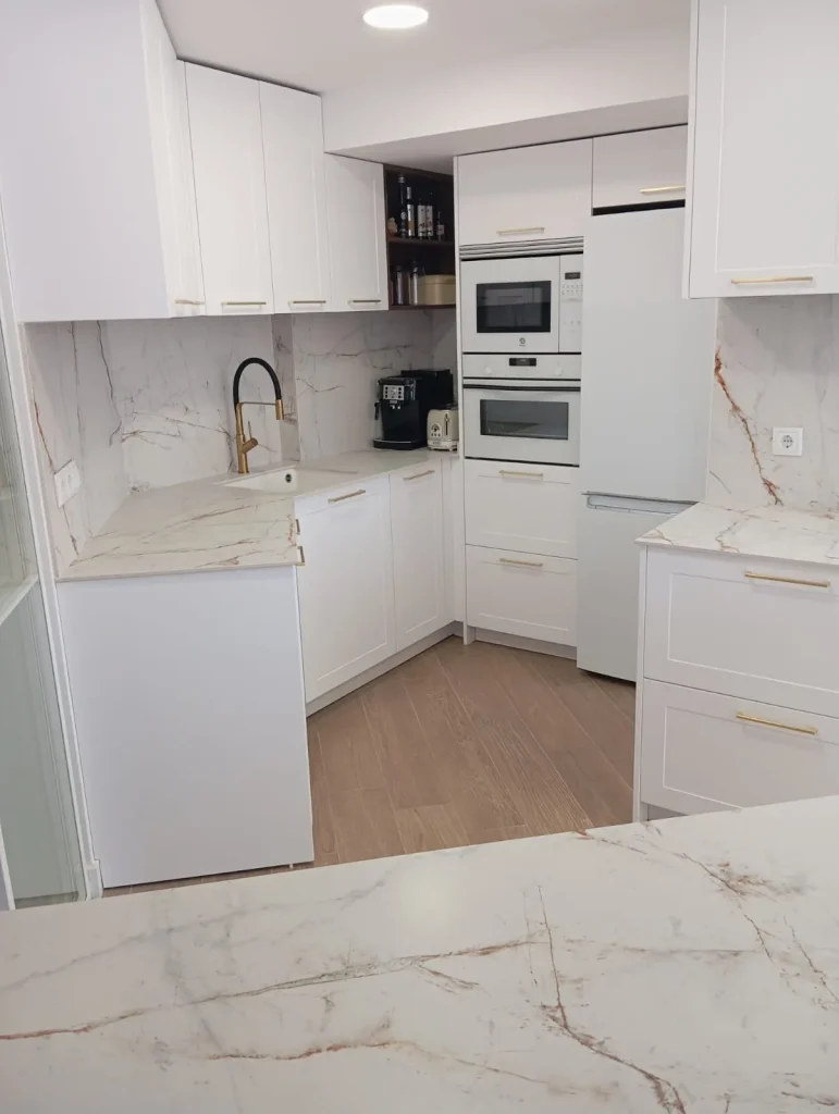 Cocina reformada en blanco con tiradores dorados, encimera efecto mármol y columna de hornos y frigorífico, en un piso de Móstoles