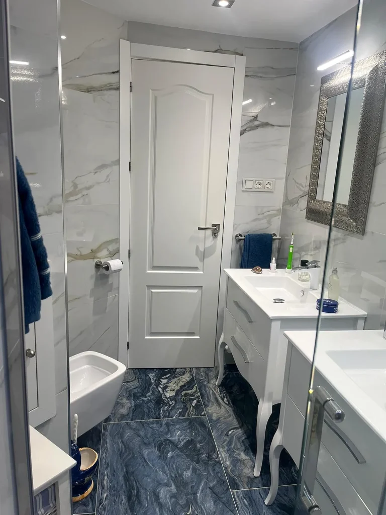 Baño reformado con revestimiento efecto mármol blanco, suelo azul veteado y mueble de lavabo blanco